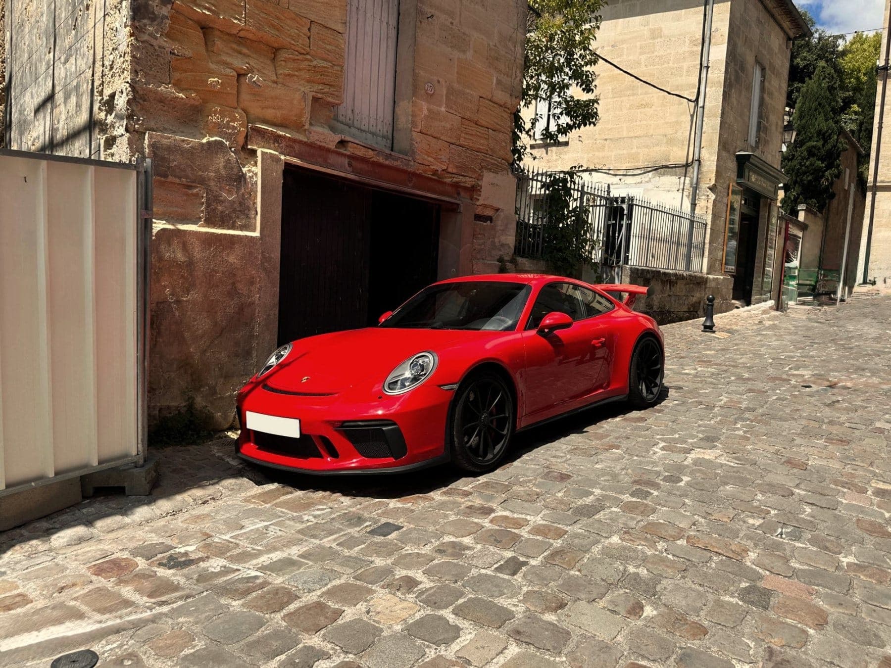 A red Porsche 911 GT3 parked on a cobbled street in an old French stone town, a garage door and shuttered houses behind it