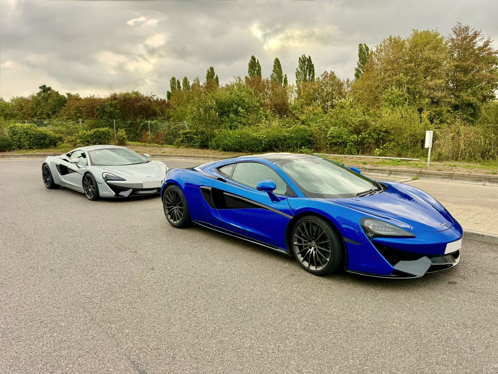 A blue McLaren 570S and a silver McLaren parked on an empty road under an autumn sky, trees and poplars behind
