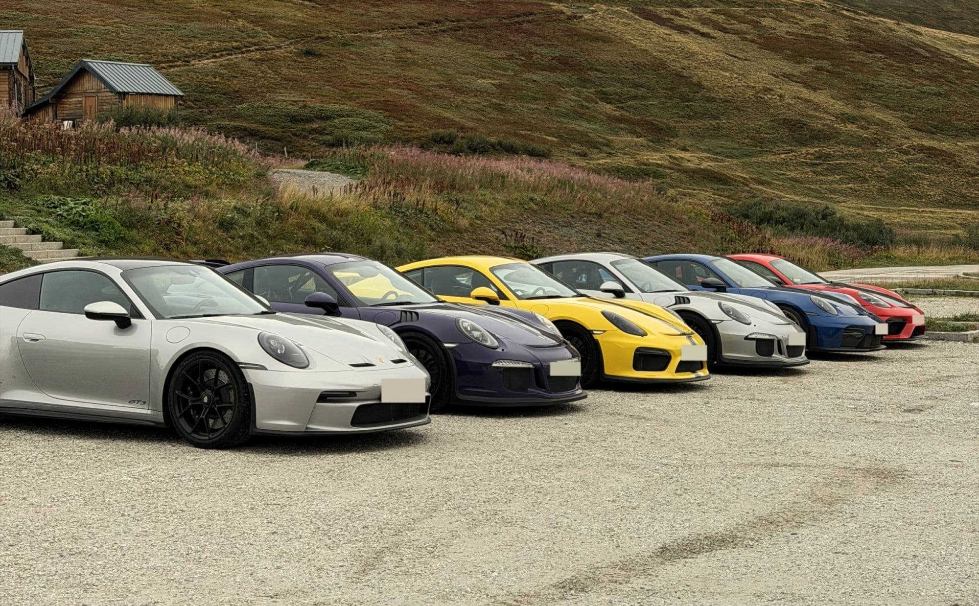 Six Porsches lined up on the gravel at the top of a mountain pass, heather and a wooden hut on the slope behind
