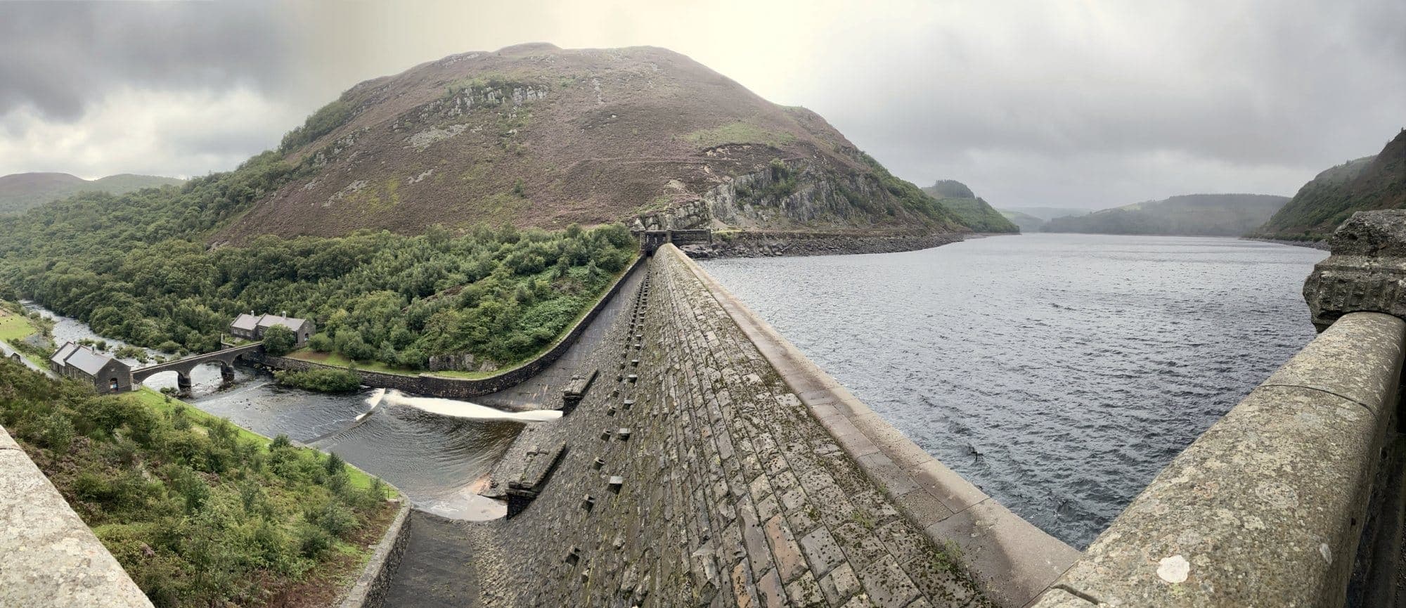 The Elan Valley above Rhayader from the top of a stone dam, the reservoir on one side and the river and an arched bridge far below on the other