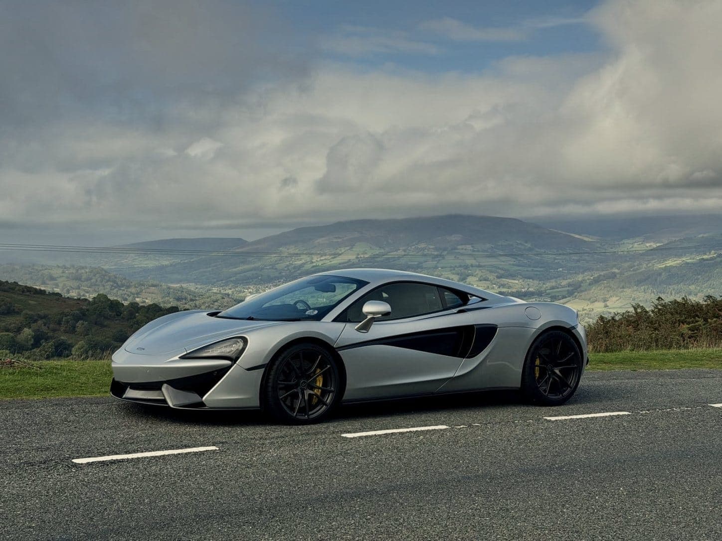 A silver McLaren with black wheels and yellow brake callipers parked on a hill road, a green valley and cloud-capped hills behind