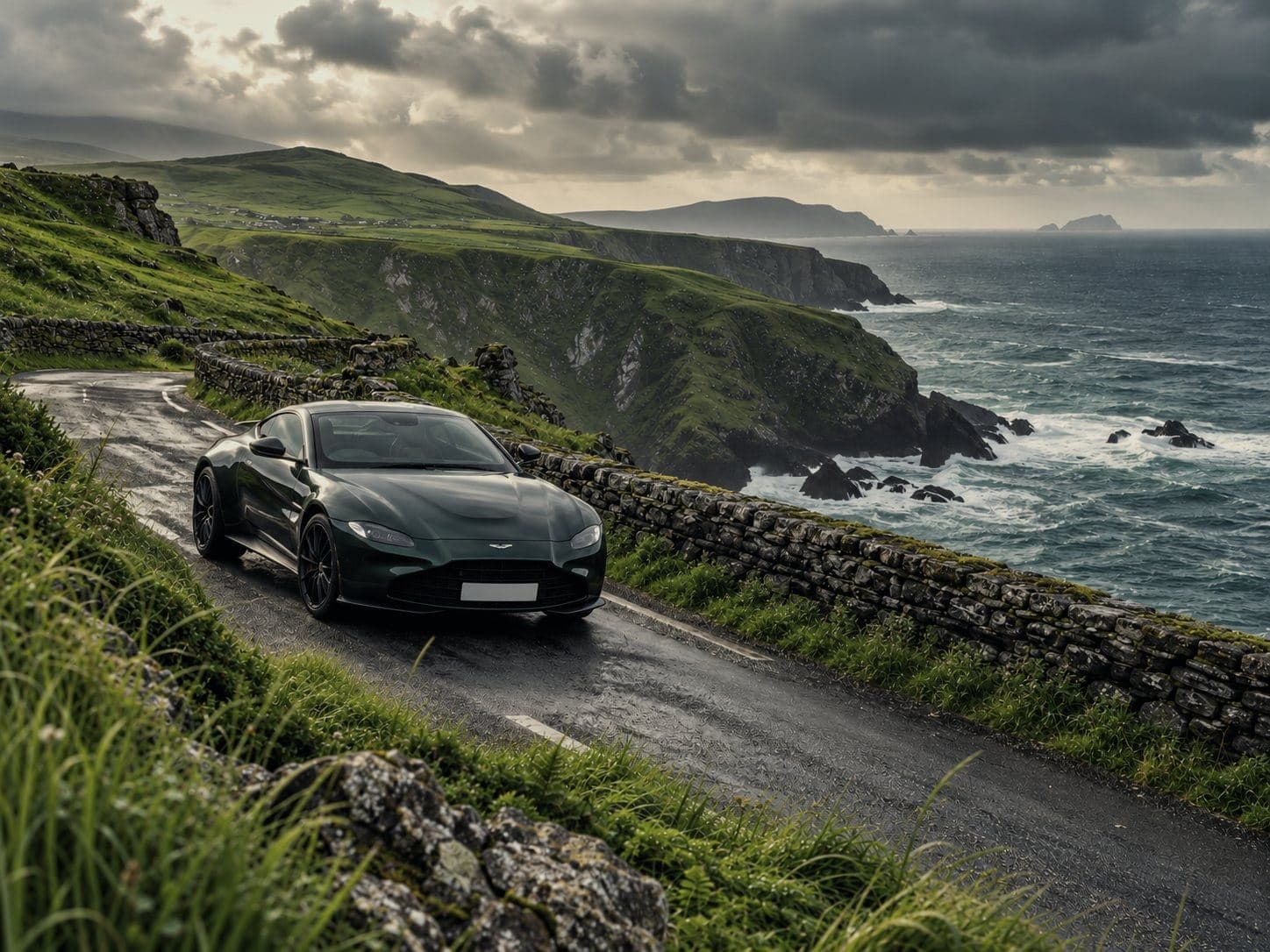 A dark green Aston Martin on a narrow coast road above Atlantic cliffs, a stone wall beside it and the sea breaking below