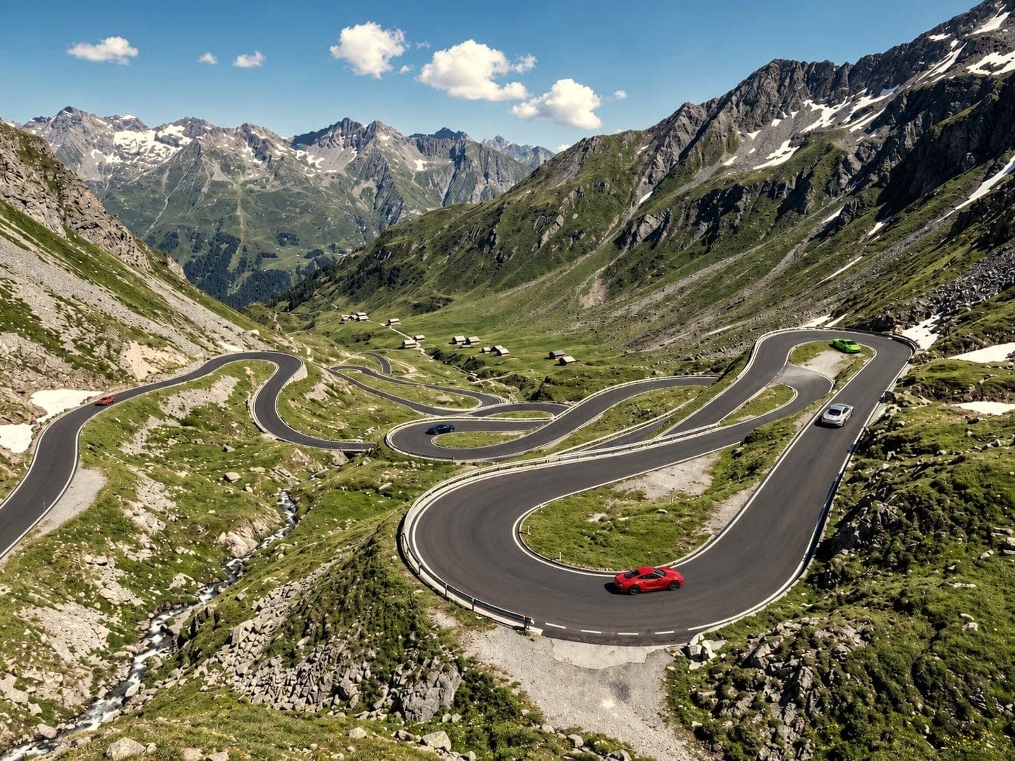 Sports cars climbing the hairpins of an alpine pass road, snow on the peaks and a stream in the valley below