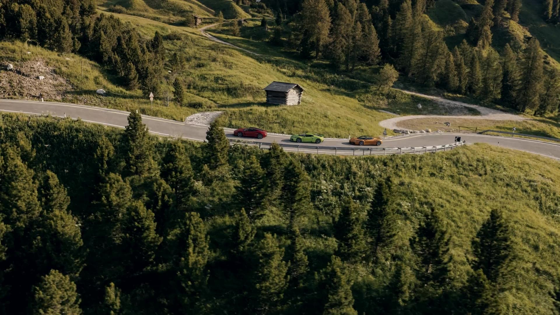 Three sports cars, red, green and orange, on a mountain pass road between alpine meadows and pine forest in the evening sun, seen from above