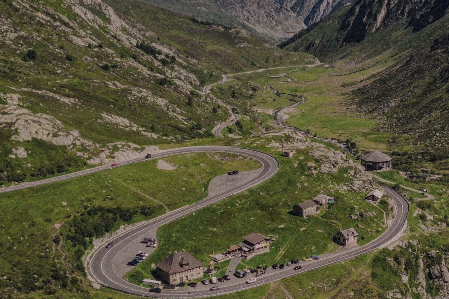 A mountain pass road doubling back on itself through a green Alpine valley, a stone inn at the hairpin