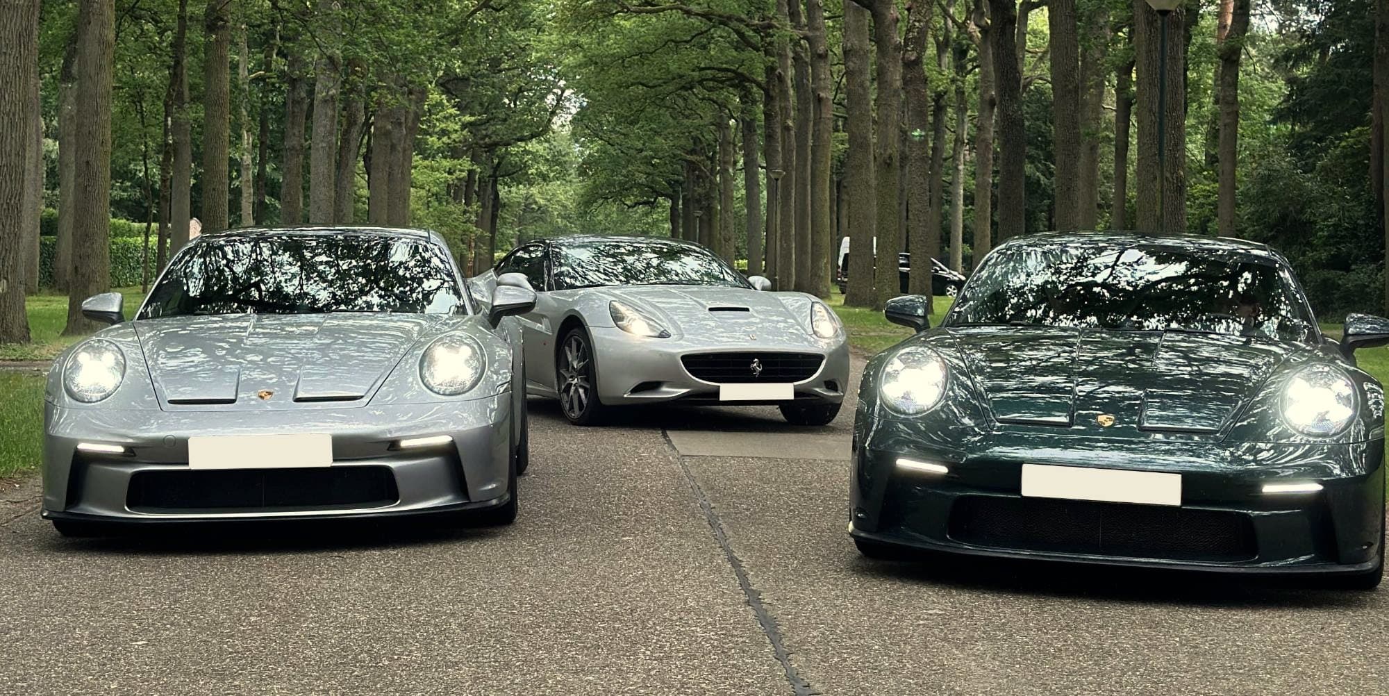 A silver Porsche 911 GT3, a silver Ferrari California and a green Porsche 911 GT3 side by side on an avenue under tall trees, headlights on