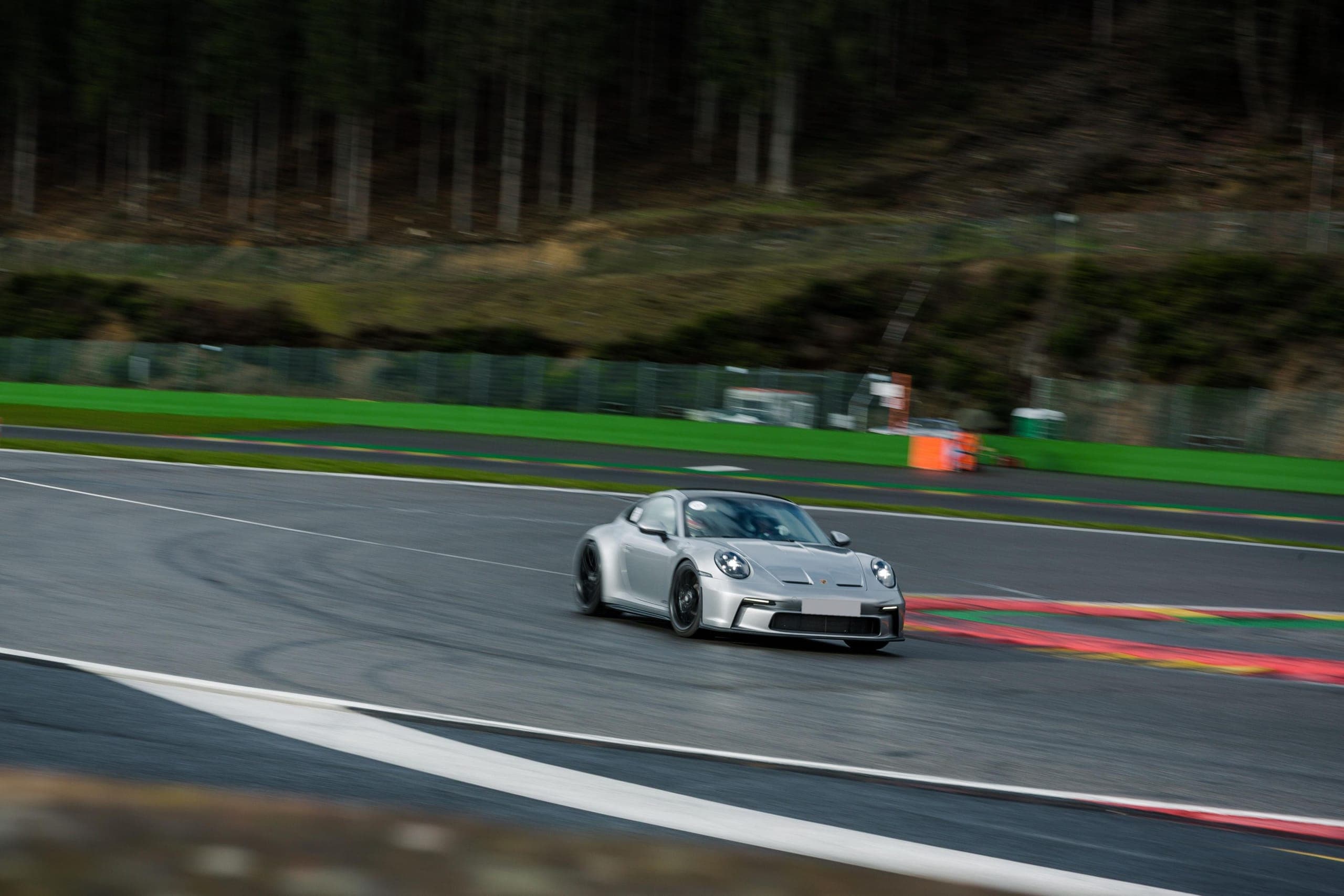 A silver Porsche 911 GT3 cornering at speed on a circuit, forest on the hillside behind and coloured kerbs at the edge of the track