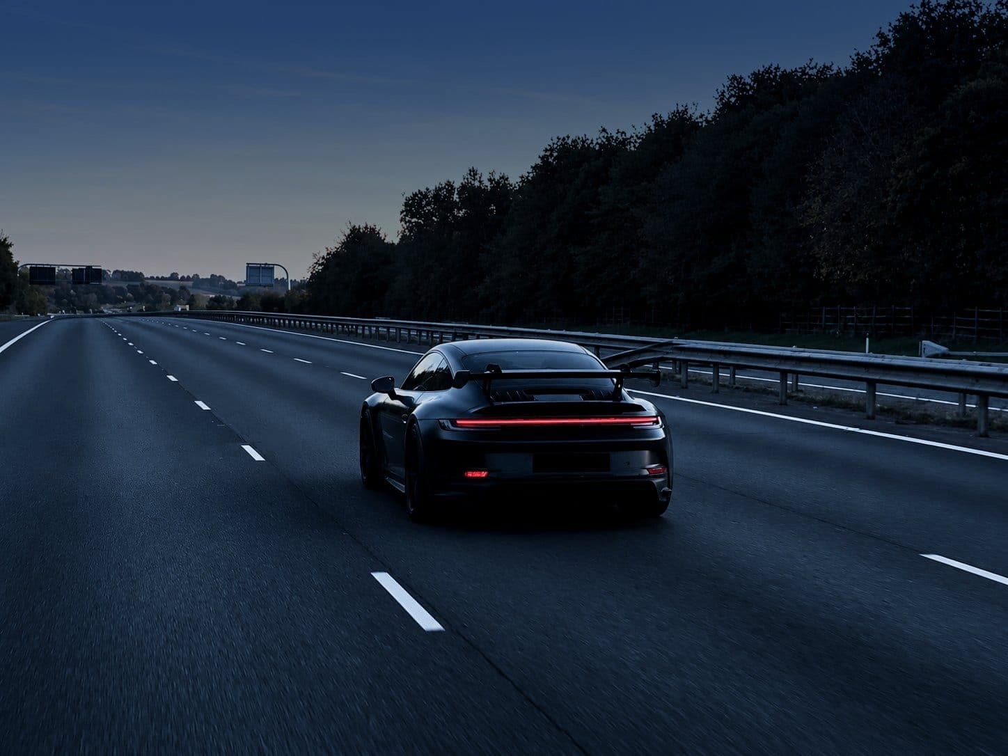 A Porsche 911 GT3 seen from behind on an empty road at dusk, its tail lights lit, trees along the verge