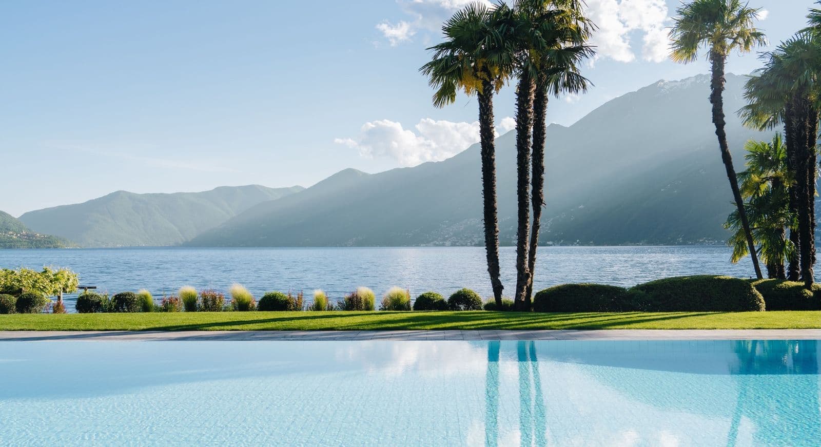 An infinity pool on a lawn beside Lake Maggiore, palms reflected in the water and the mountains beyond in the afternoon light