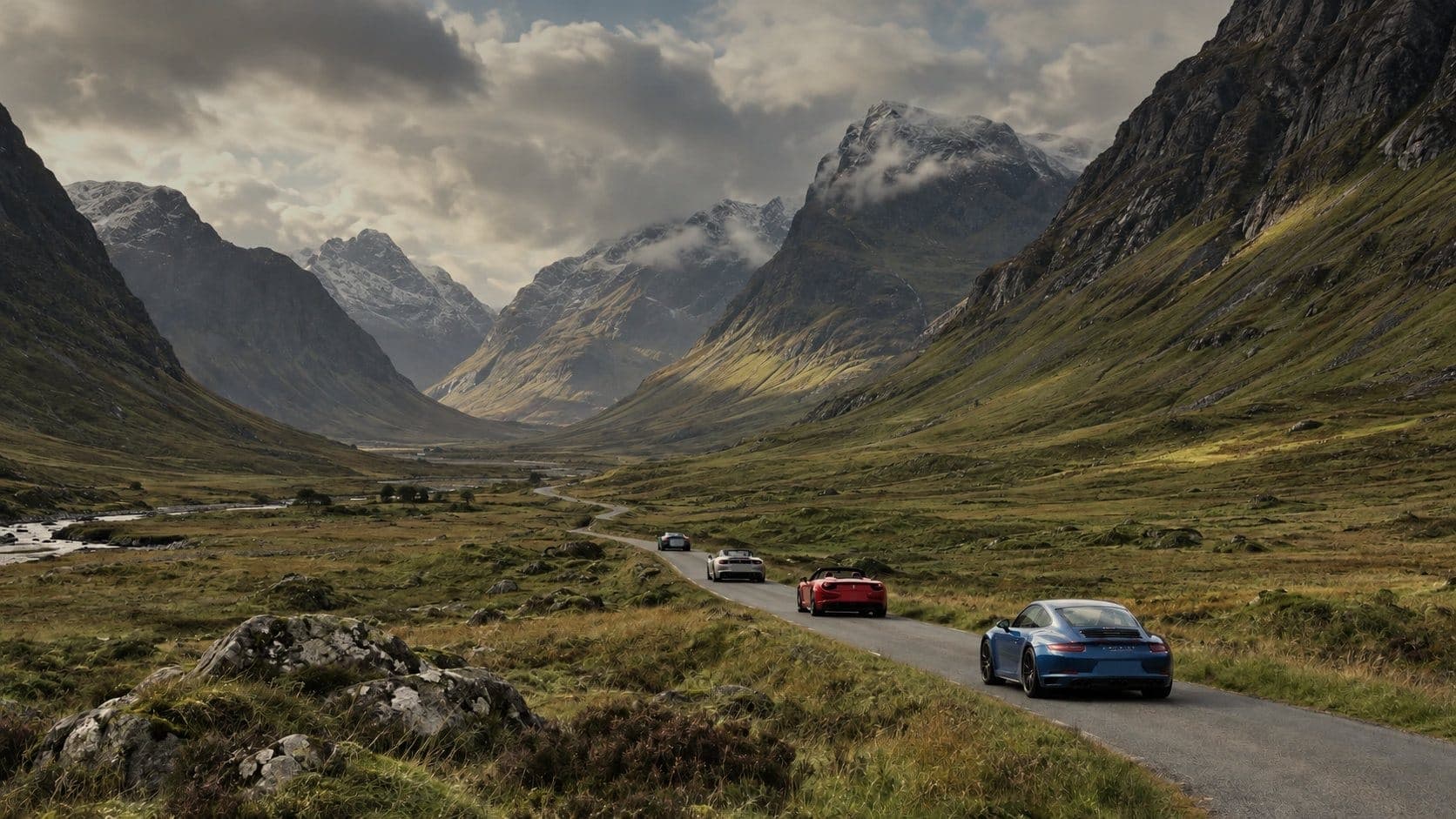 Four sports cars in a line on a single track road through a Highland glen, snow on the peaks and cloud on the tops